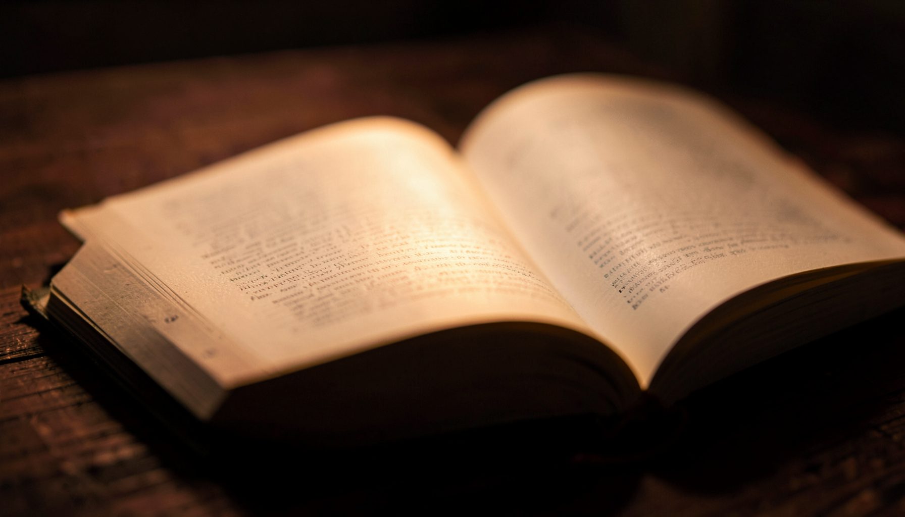 A moody, warm-toned close-up of an open hardcover book resting on a dark wooden table, with soft amber light spilling across the pages and deep shadows pooling around the edges.