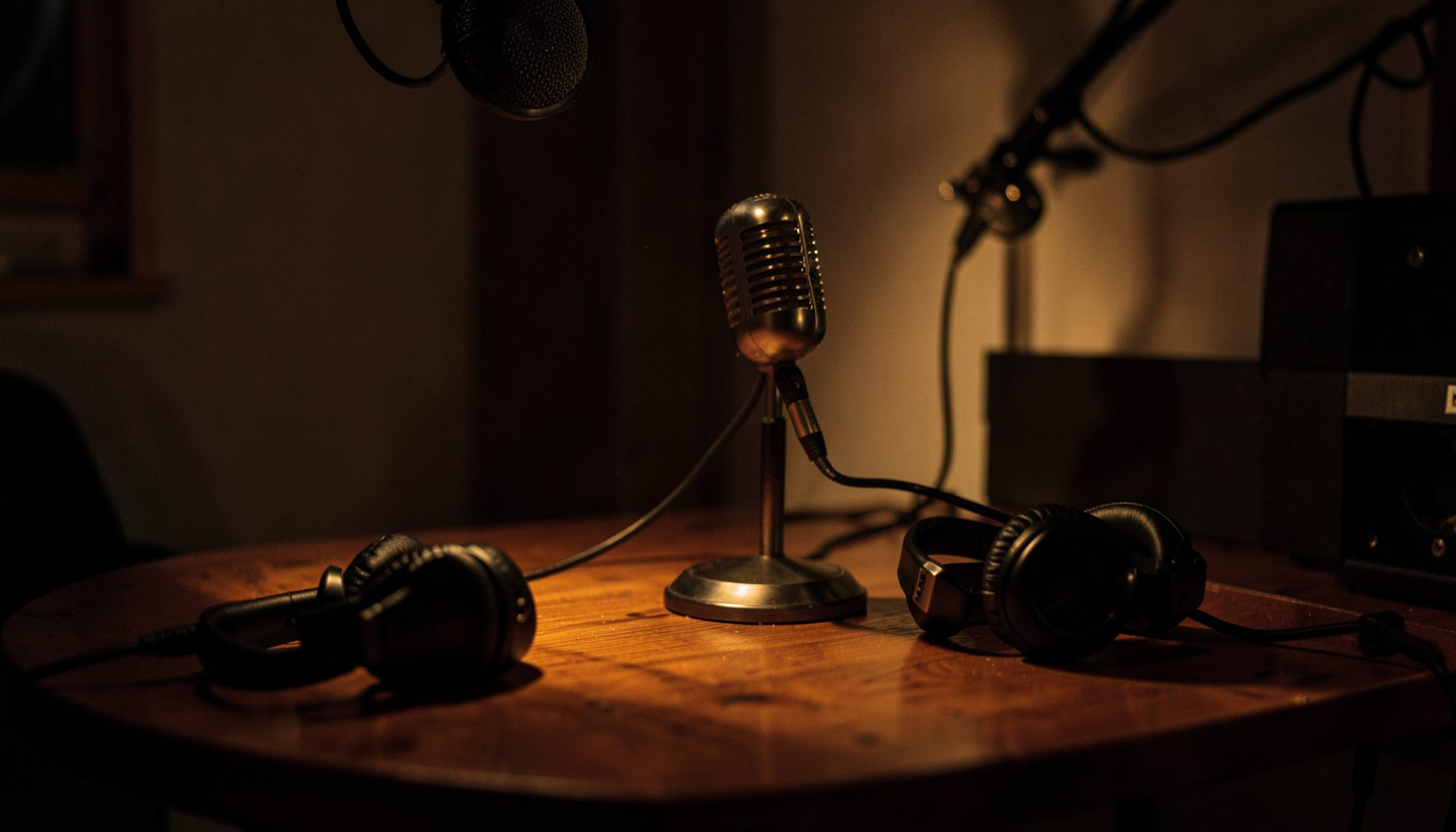 A warm, dimly lit podcast recording setup with a vintage microphone on a wooden desk, headphones draped nearby, and a soft amber glow casting long shadows across the room.